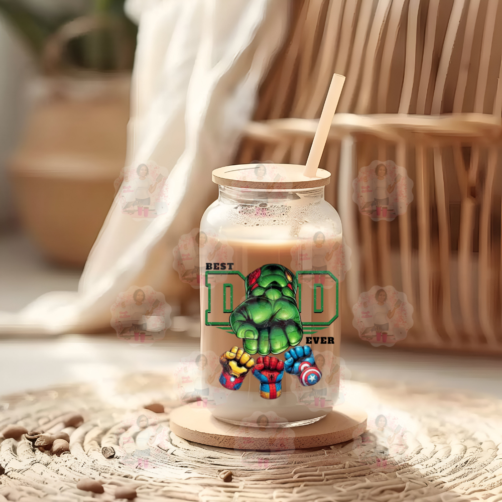 A glass tumbler with a 'Best Dad Ever' graphic featuring a cartoonish green character resembling a well-known superhero, placed on a wicker surface with a chair and sheer curtains in the background.